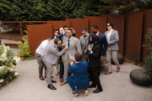 Wedding party members in gray suits and blue dresses gather on a stone patio during an outdoor celebration.