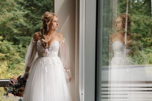 A bride in a flowing white tulle gown stands near a window in natural light.