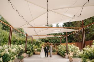 An outdoor wedding ceremony setup with white canopy sails and lush floral arrangements in a garden setting.