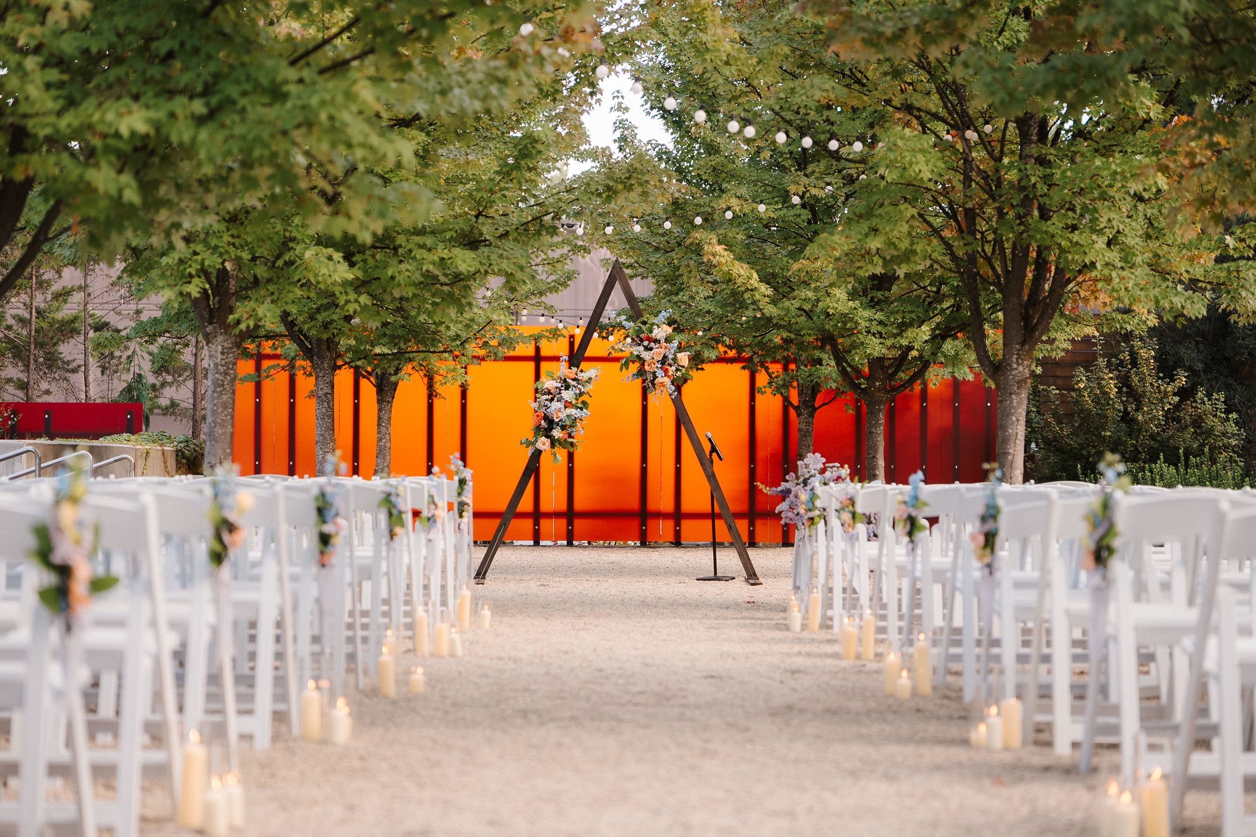 White chairs line a path for an outdoor wedding ceremony with orange and red decorative panels in the background.