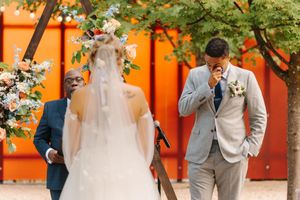 A bride and groom share romantic moments in front of a vibrant orange wall with lush greenery.