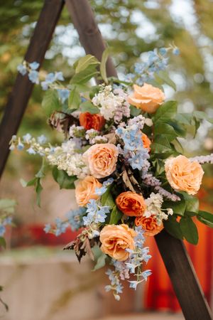 A romantic floral arrangement with peach roses and light blue delphinium flowers hanging on a wooden arch outdoors.