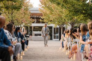 Wedding guests gather outdoors in a courtyard surrounded by autumn trees during a sunny afternoon celebration.