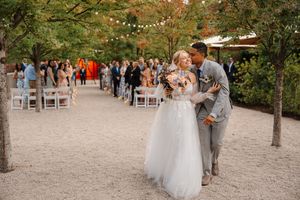 A rustic outdoor wedding ceremony setup with white chairs arranged on a gravel path surrounded by trees and greenery.