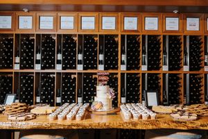 Wedding cake display with wine bottles and desserts arranged on wooden tables in a rustic venue.