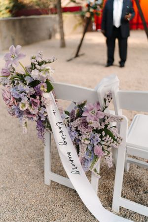 White wedding ceremony chair decorated with purple and pink floral arrangements and a white ribbon at an outdoor venue.