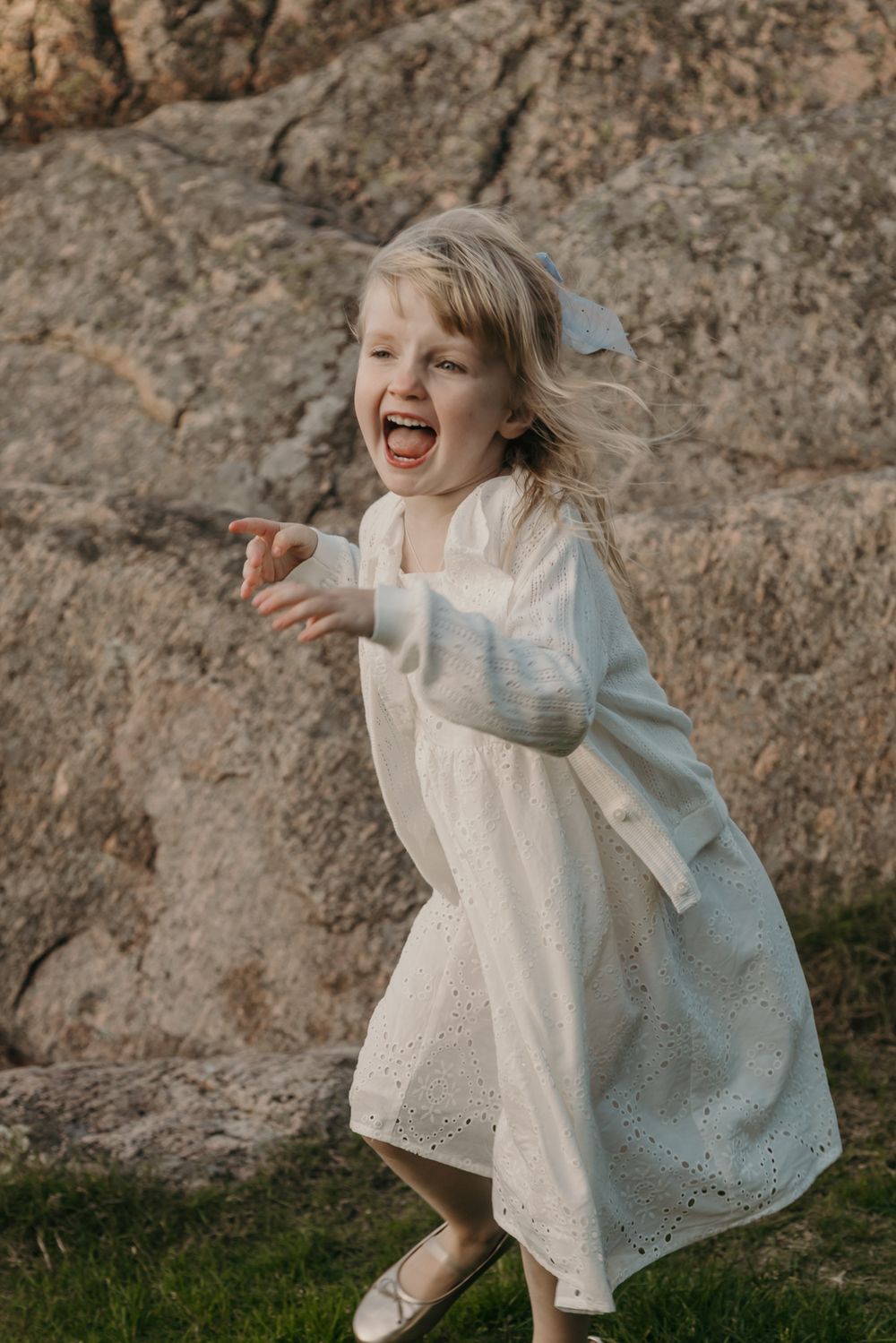 A joyful child in a white eyelet dress dances and plays against a rocky outdoor backdrop.