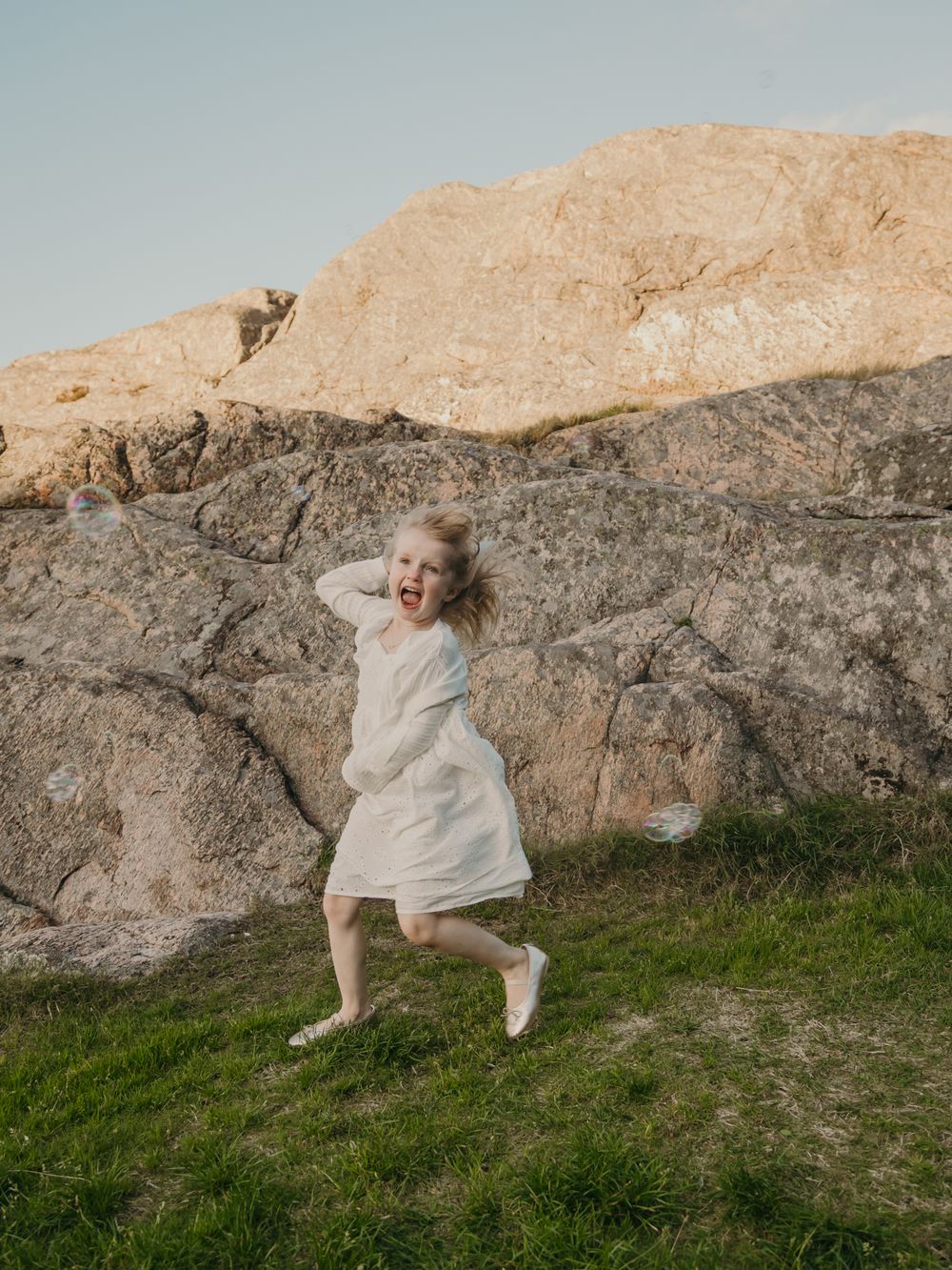 A figure in a flowing white dress moves gracefully against a rocky cliff backdrop with green grass below.