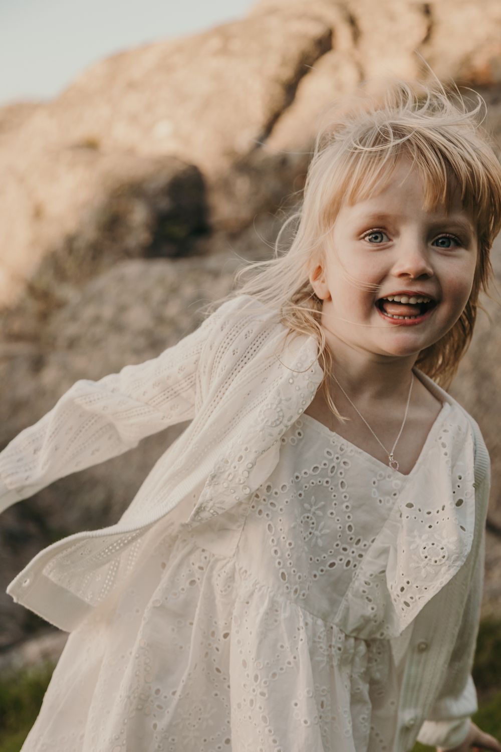 A candid portrait of pure childhood joy captured in white lace dress against natural rock formations.
