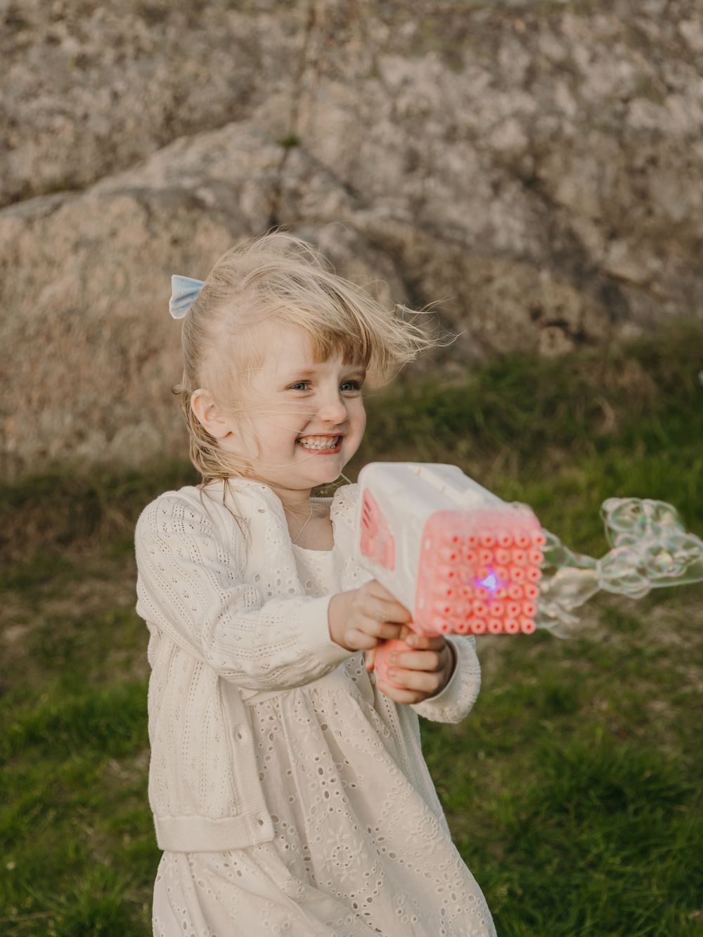 A child in a white eyelet dress holds and plays with a pink polka dot gift box against a stone wall background.