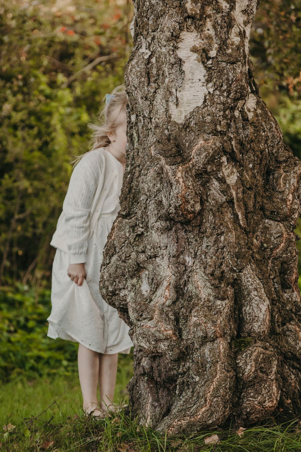 A person in a white dress stands behind a large tree trunk with textured bark in a natural outdoor setting.
