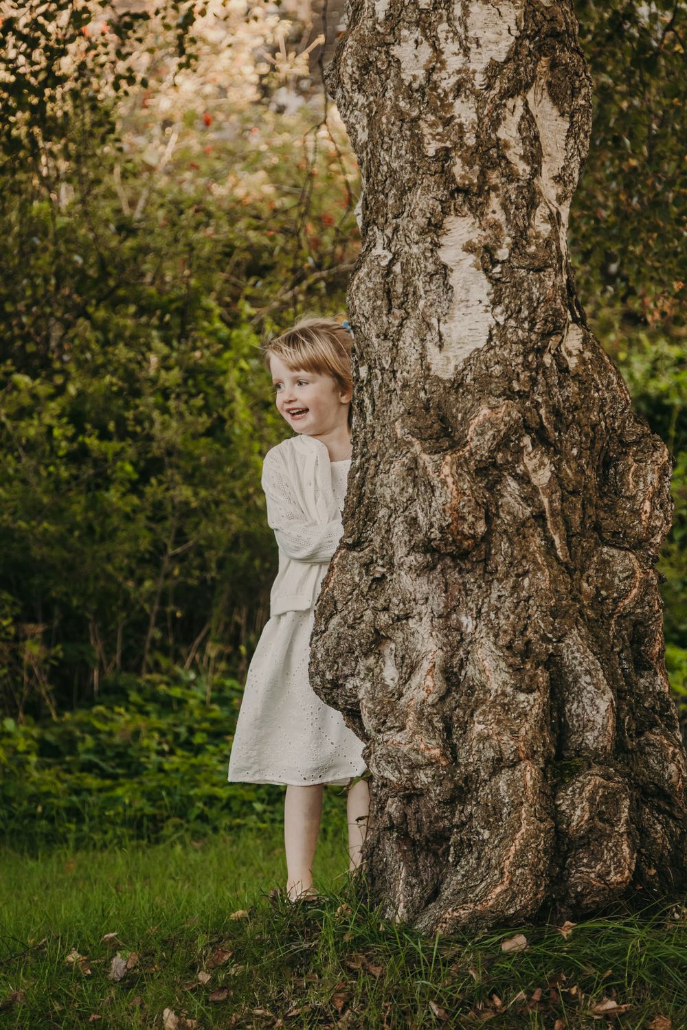 A child in a white dress peeks playfully from behind a large tree trunk in a lush green outdoor setting.