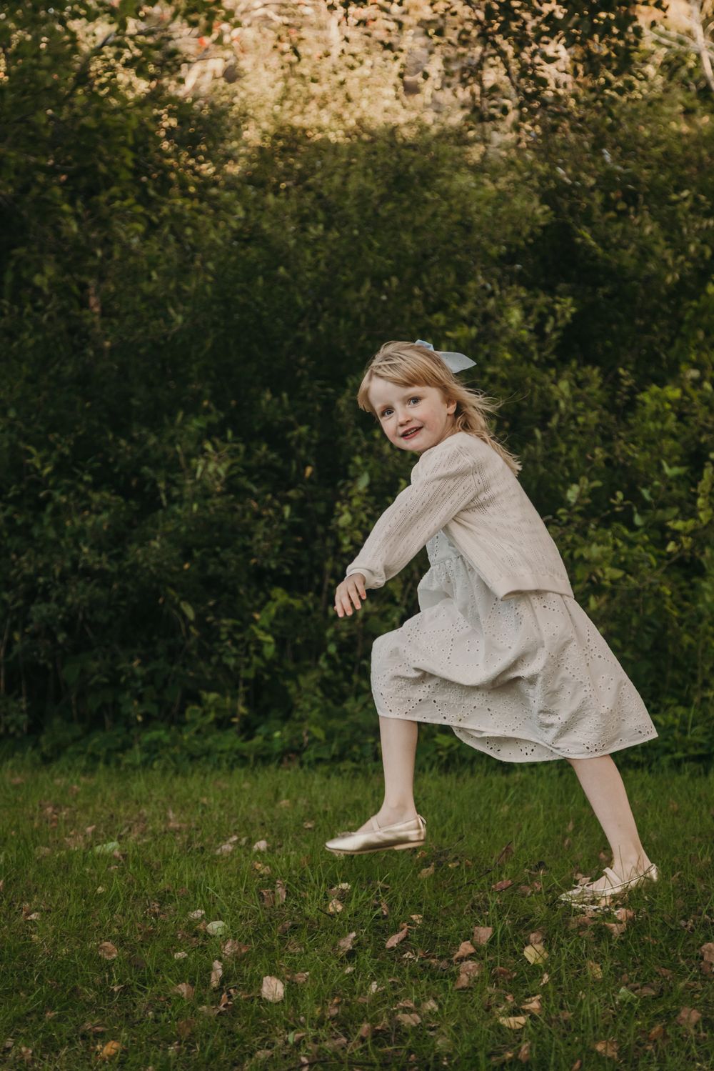 A child in a flowing dress dances and twirls in a sunlit garden with fallen leaves scattered on the grass.