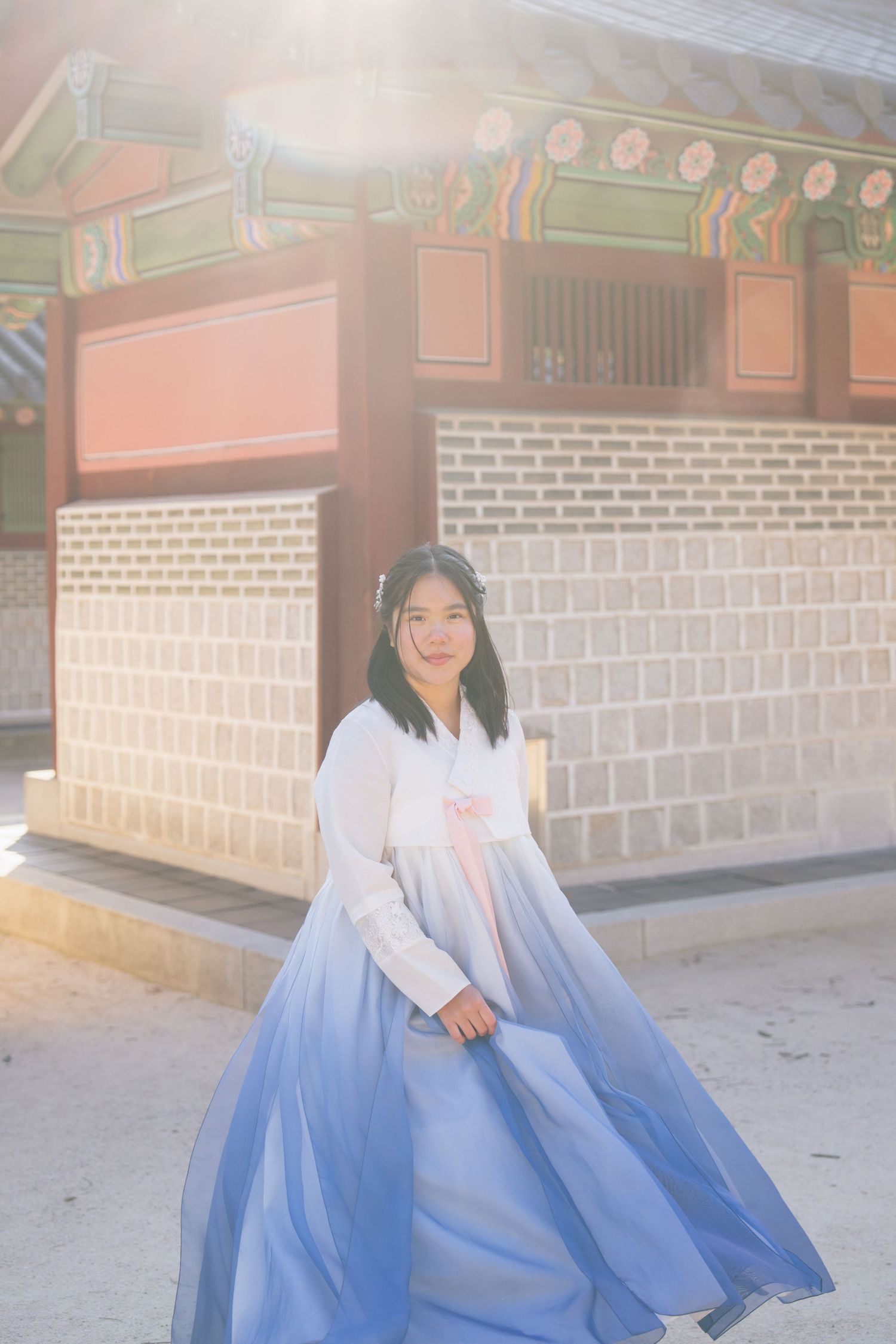 A person wearing a traditional Korean hanbok dress in white and blue sits in front of a temple wall.