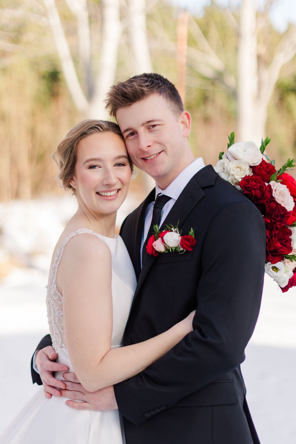 A romantic winter wedding couple poses together in formal attire with red and white roses against a snowy backdrop.