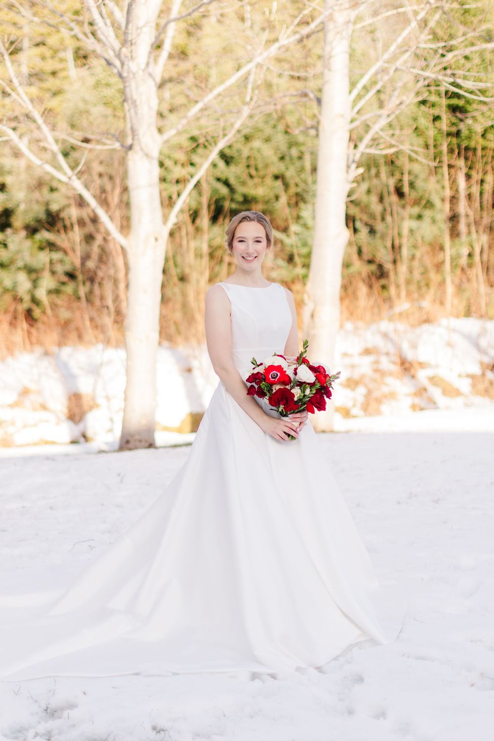 A bride in a white gown holds a vibrant red bouquet while standing in snow with bare trees in the background.