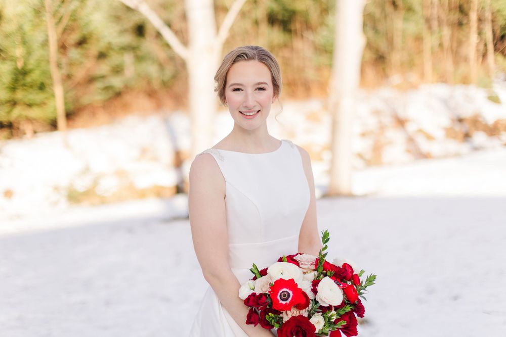 Winter bride in white dress holds vibrant red and white bouquet against snowy outdoor backdrop.