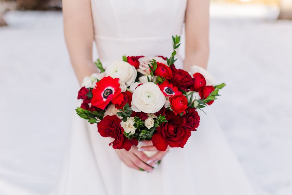 A stunning bridal bouquet featuring vibrant red anemones, roses and white ranunculus flowers against a white background.