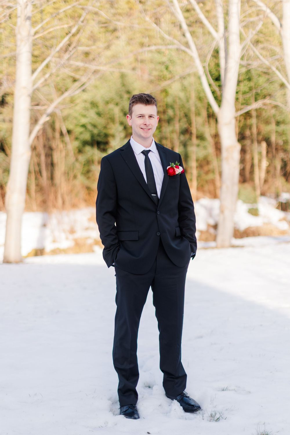 A person in a black suit with a red boutonniere stands in a snowy outdoor setting with white birch trees in the background.