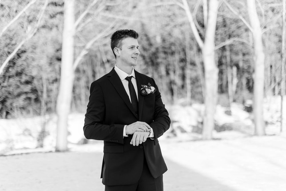 A groom in a black suit laughs and dances outdoors in a winter setting with snow-covered trees in the background.