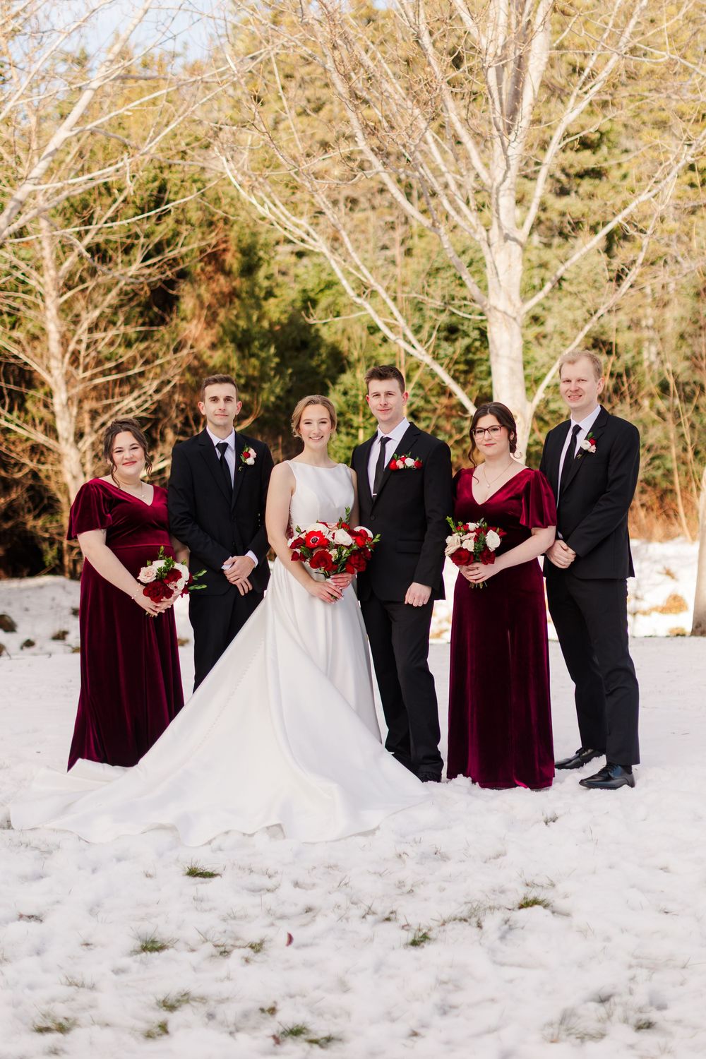 Wedding group stands in snow with bare trees, featuring burgundy dresses and black suits.