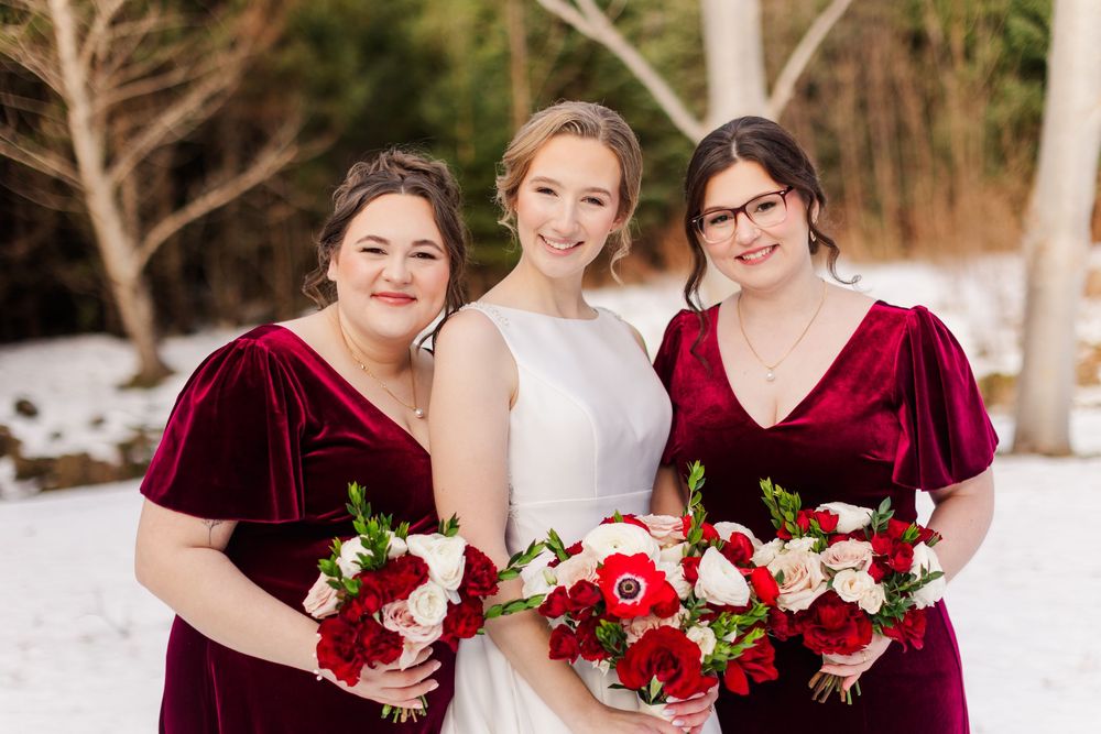Bridal party laughs together in snow with burgundy bridesmaid dresses and red bouquets.