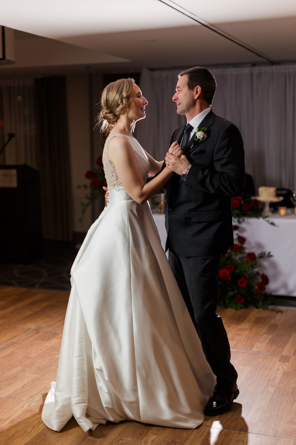 A couple in formal wedding attire share an intimate dance moment on a wooden floor with red floral decor in the background.