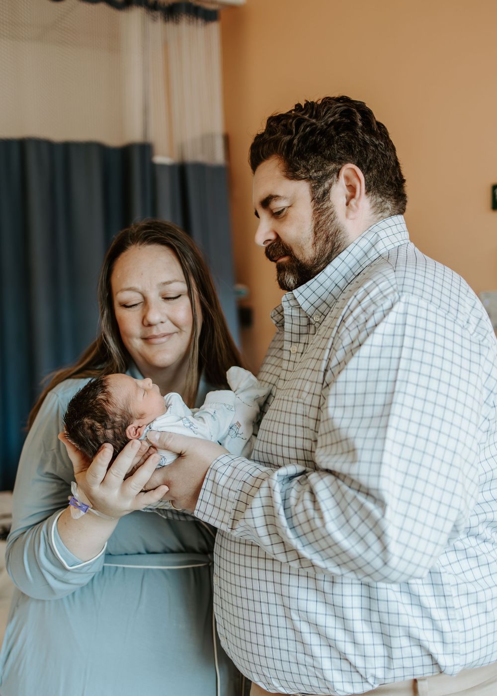 A couple smiles lovingly while holding their newborn baby in a hospital room.