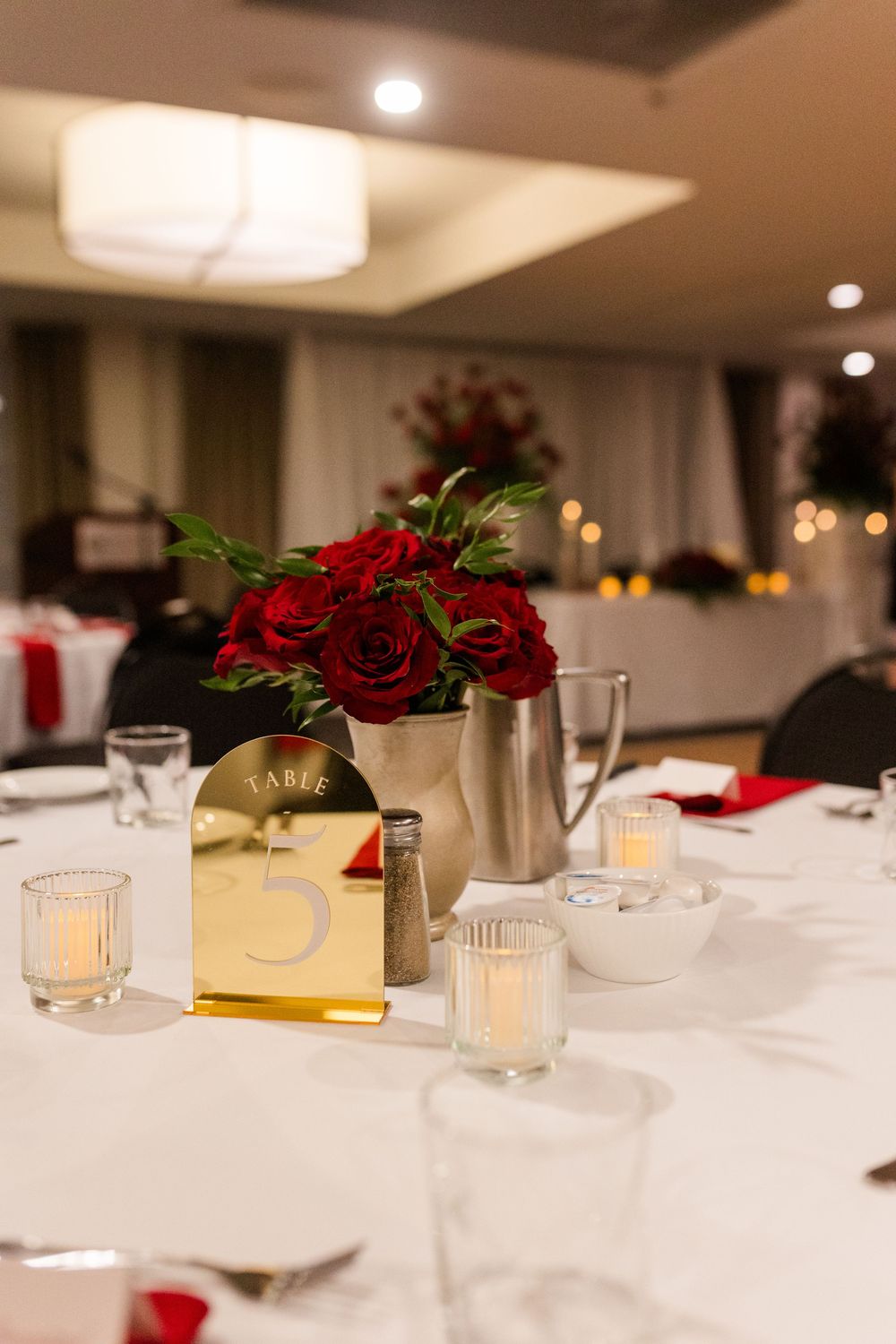 Elegant table setting with red rose centerpieces and glowing candles on white tablecloths in a banquet hall.