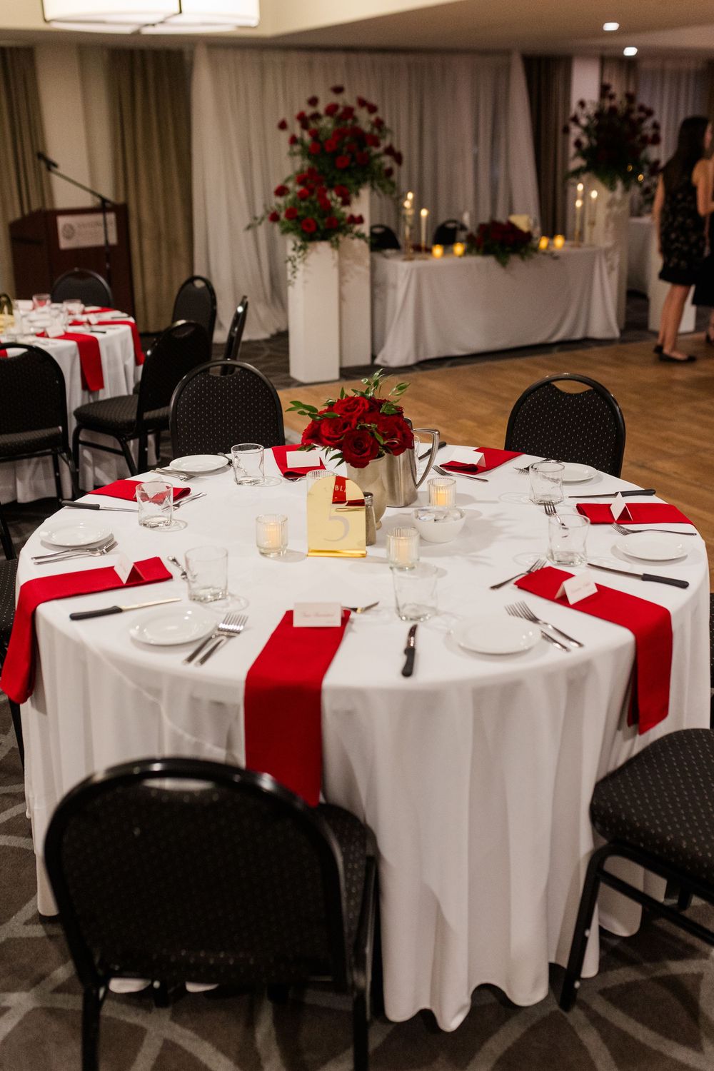 Elegant wedding reception table with white linens, red runners, and floral centerpieces in a banquet hall setting.
