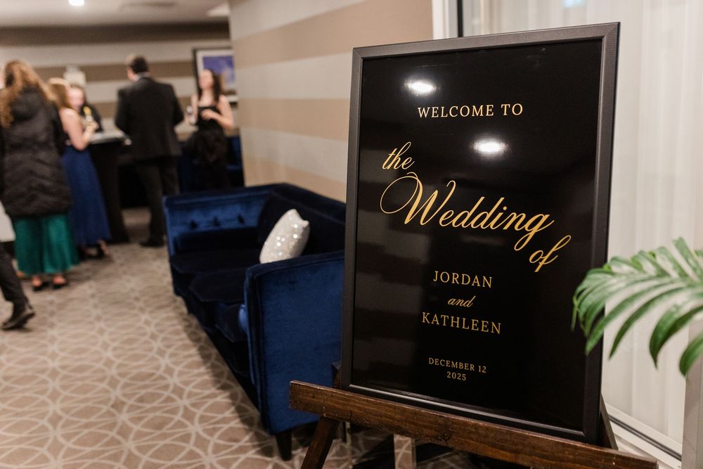 A black and gold wedding welcome sign stands next to a blue velvet couch at an elegant venue entrance.