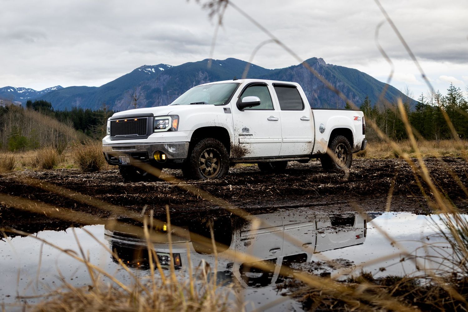 White GMC Sierra pickup truck parked near a puddle reflecting mountain scenery and cloudy skies.