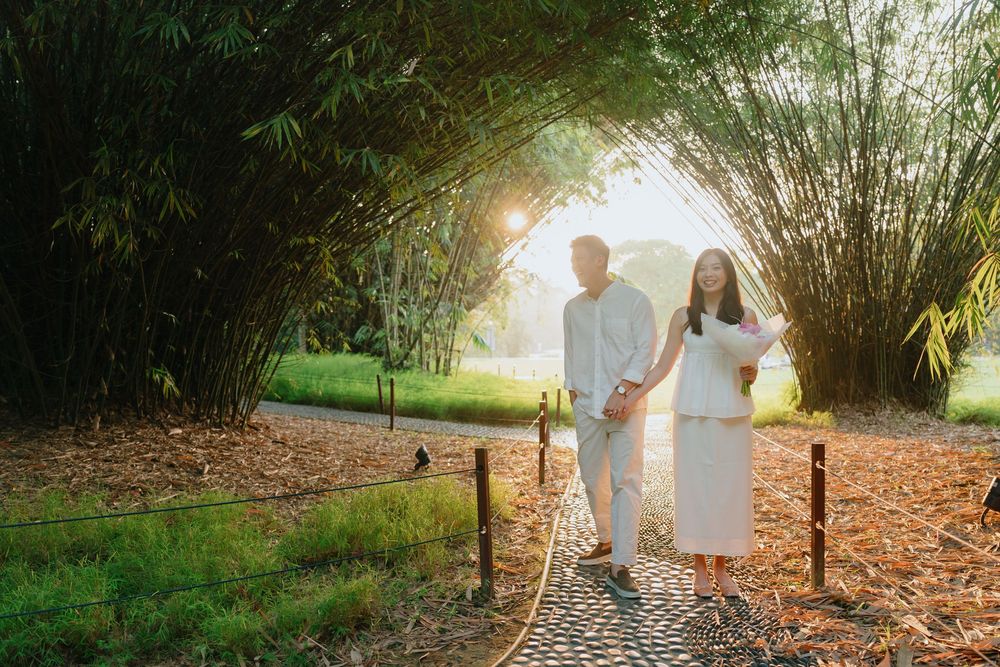 A couple in white outfits walks along a tree-lined path during an autumn engagement photo session.