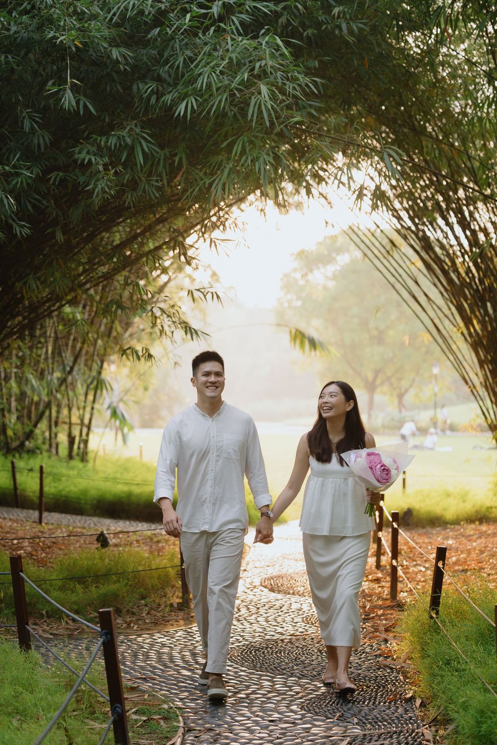 A couple walks hand in hand along a winding stone path through a lush bamboo garden at sunset.