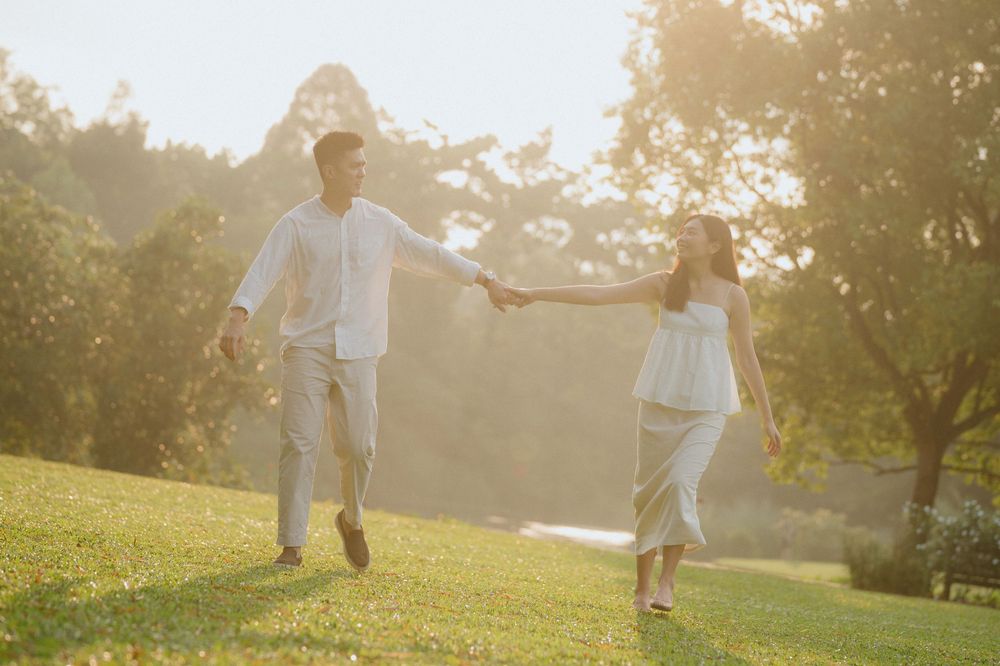 A couple in white outfits holds hands while running through a sunlit park at golden hour.