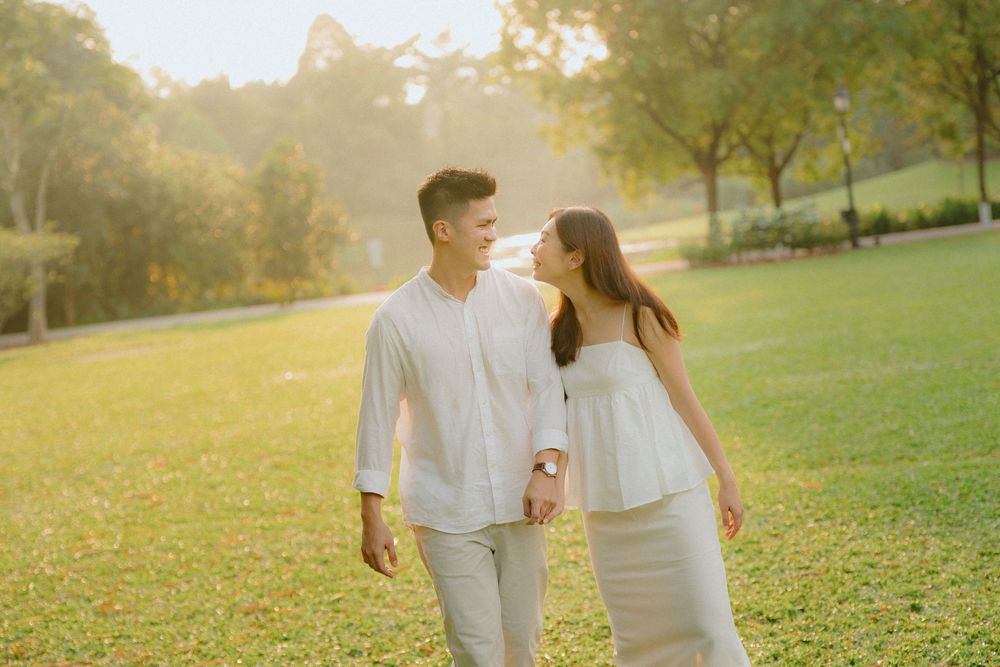 A couple in white outfits walks together through a sunlit park during golden hour.