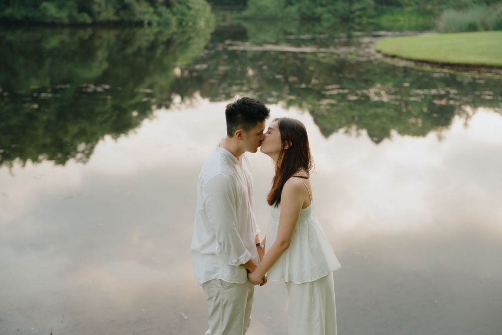 Couple in white wedding attire shares a romantic moment by a serene lake surrounded by lush greenery.