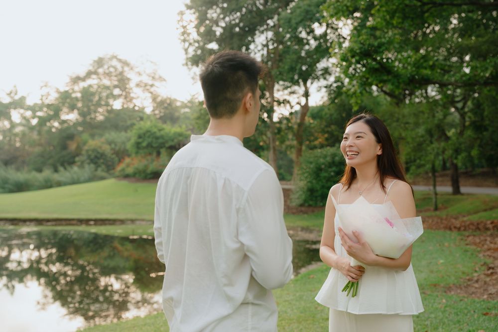 A couple in white outfits sharing a joyful moment by a serene pond in a park setting during golden hour.