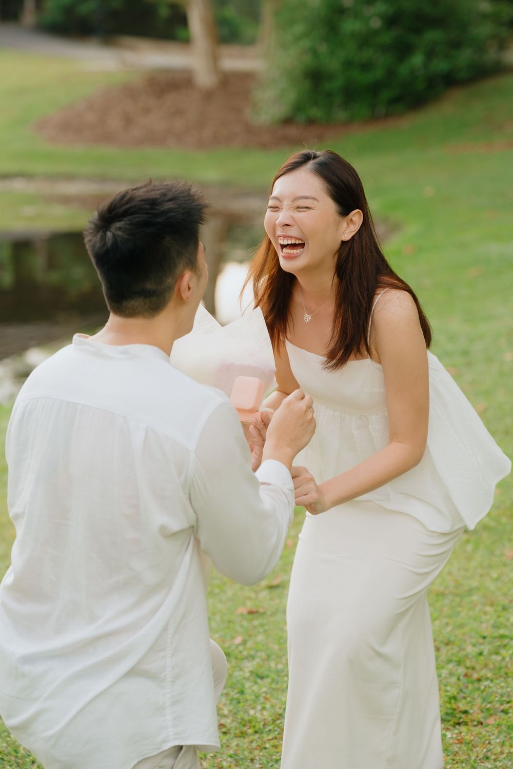 A couple in white outfits laughs and embraces joyfully in a green outdoor garden setting during a photoshoot.