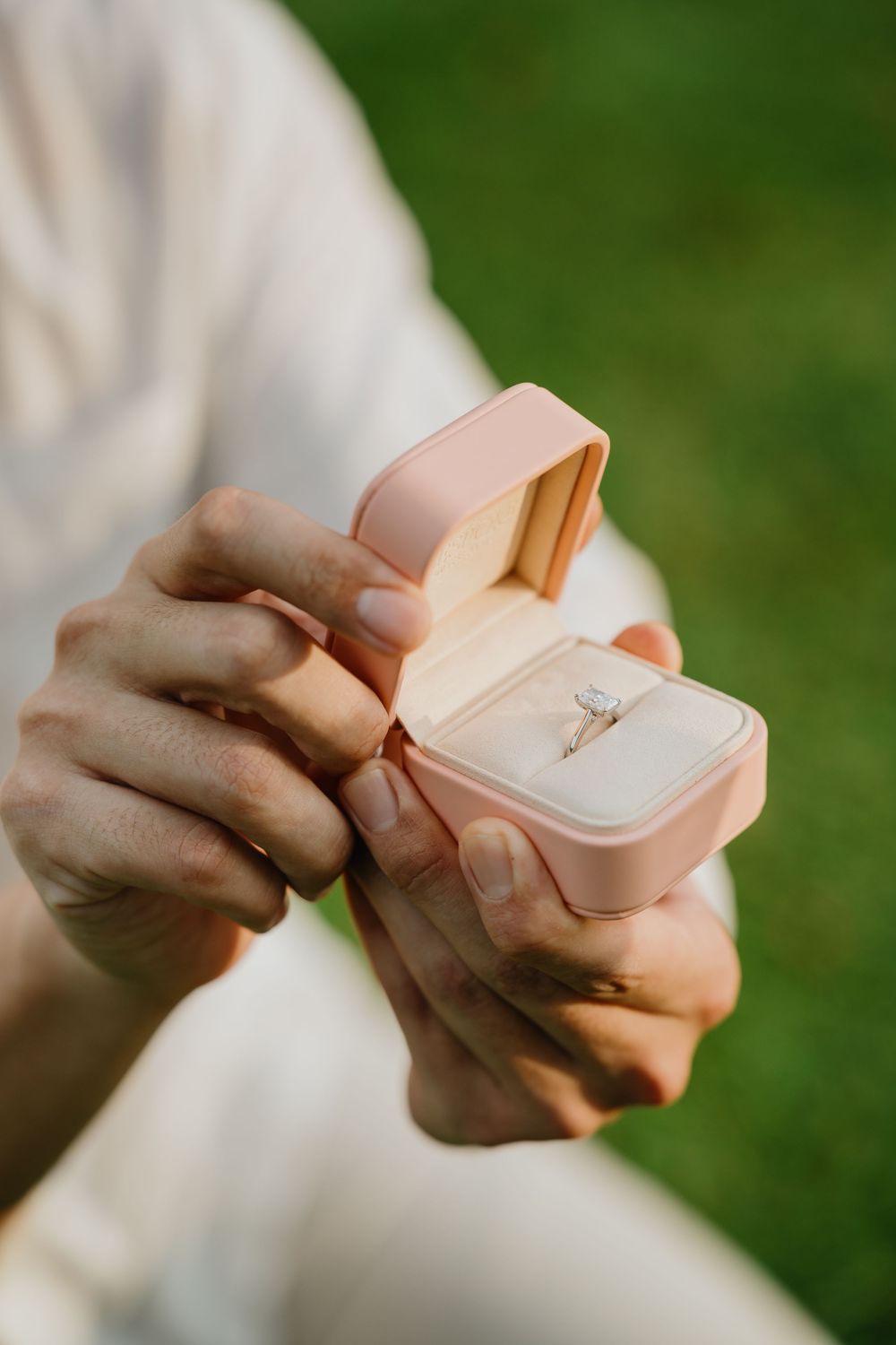 Hands holding an open blush pink engagement ring box with a diamond solitaire ring against a green outdoor background.