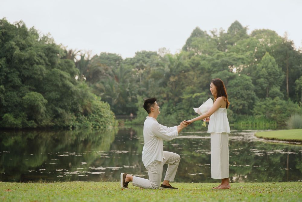 A romantic lakeside proposal scene with two people in white outfits surrounded by lush greenery and water.