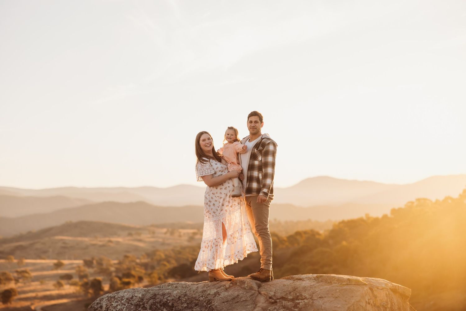 Couple embraces while standing on hilltop against golden sunset in desert landscape.