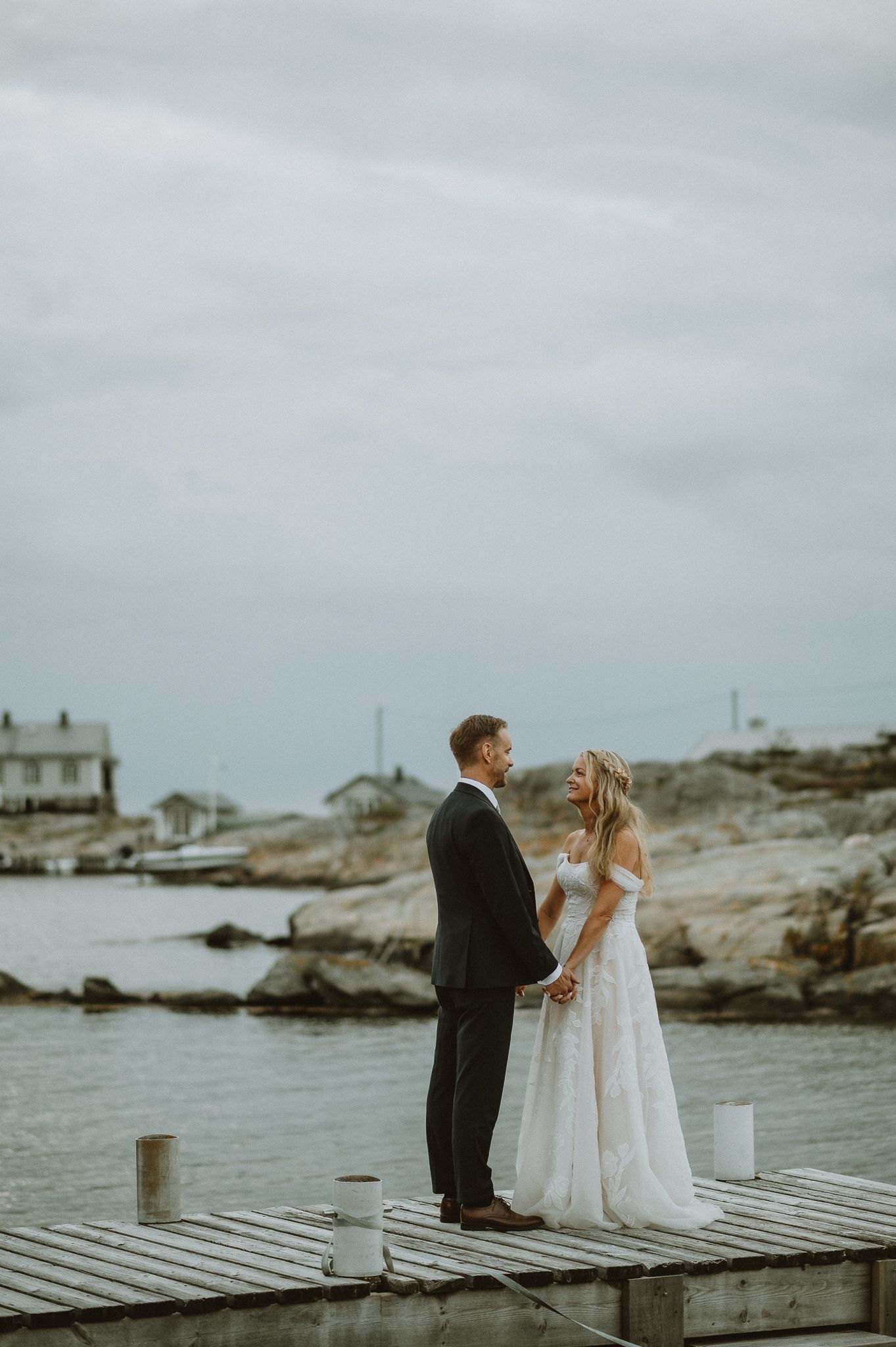 Brudpar står på en brygga vid Sillsalteriet, Marstrand och ser på varandra. Det är kvällsljus och man ser klippor och ett hus i bakgrunden.