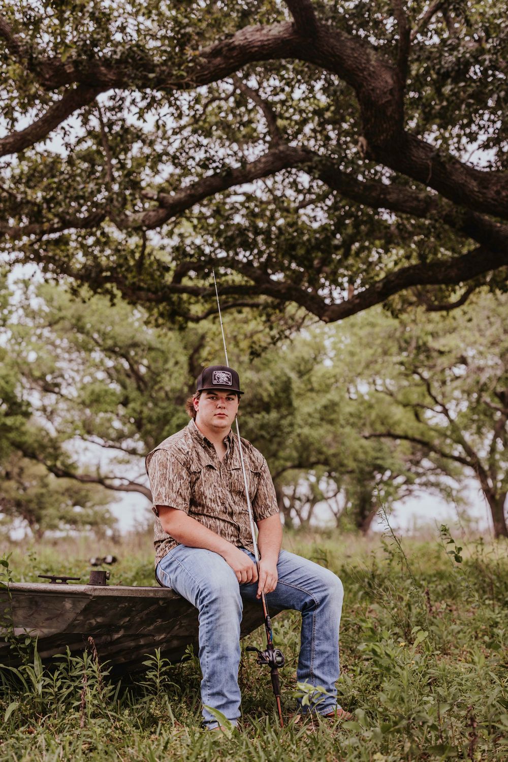 Person in camo shirt and baseball cap relaxes on wooden bench under tree branches.
