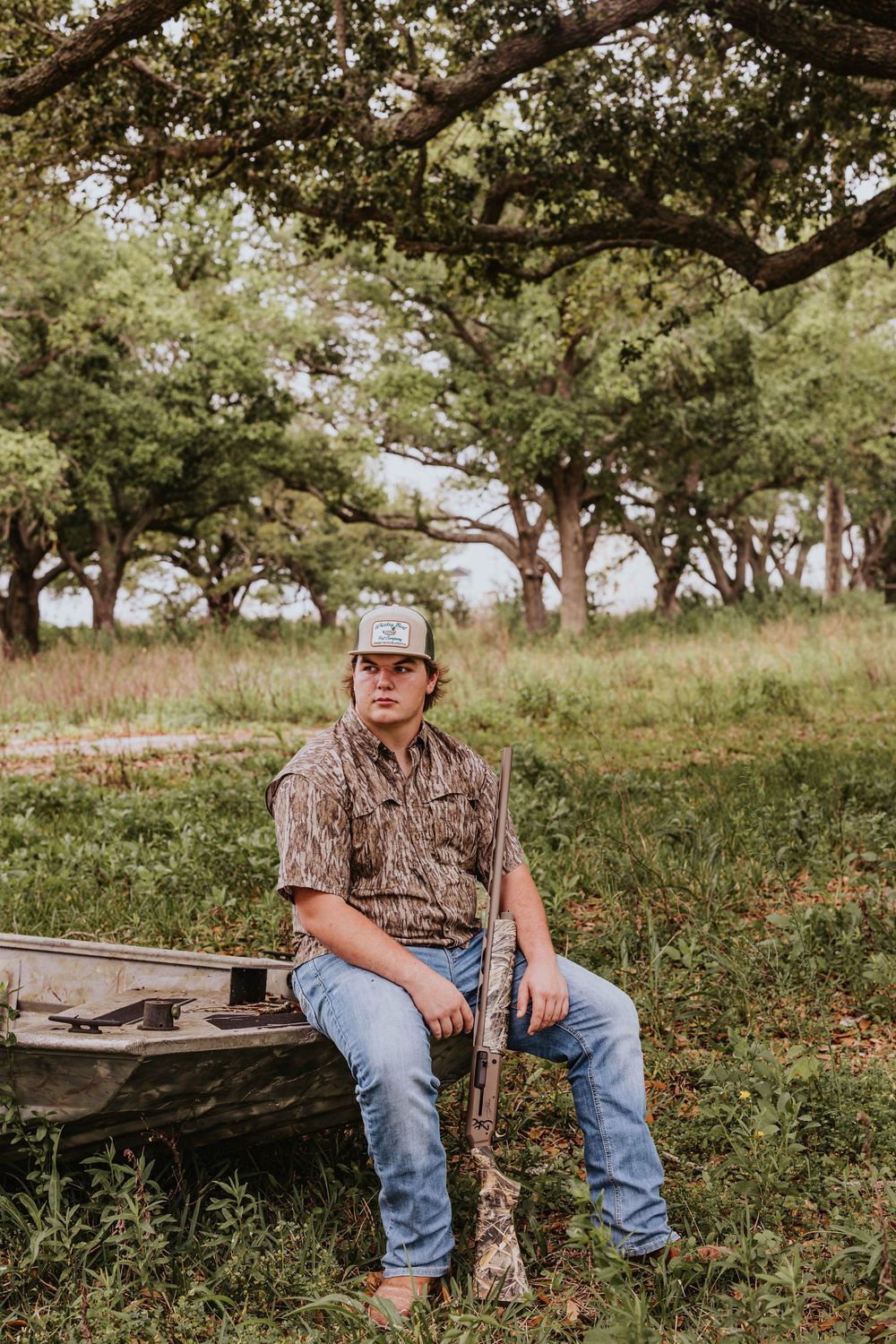 Person in hunting attire sits on wooden bench in rural landscape.