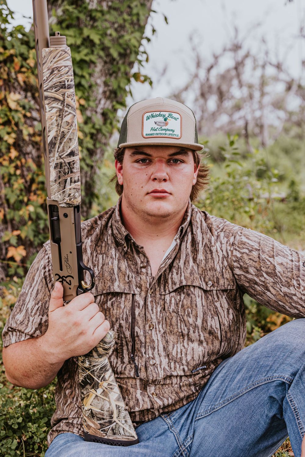 Person in camouflage clothing poses with hunting equipment in outdoor setting.