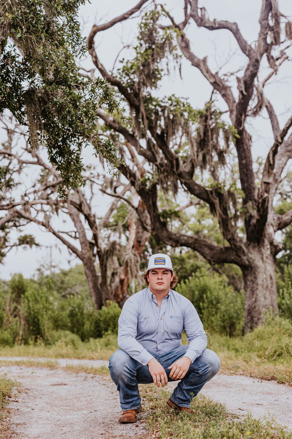 Person in baseball cap and jeans sits on ground with gnarled trees in background.
