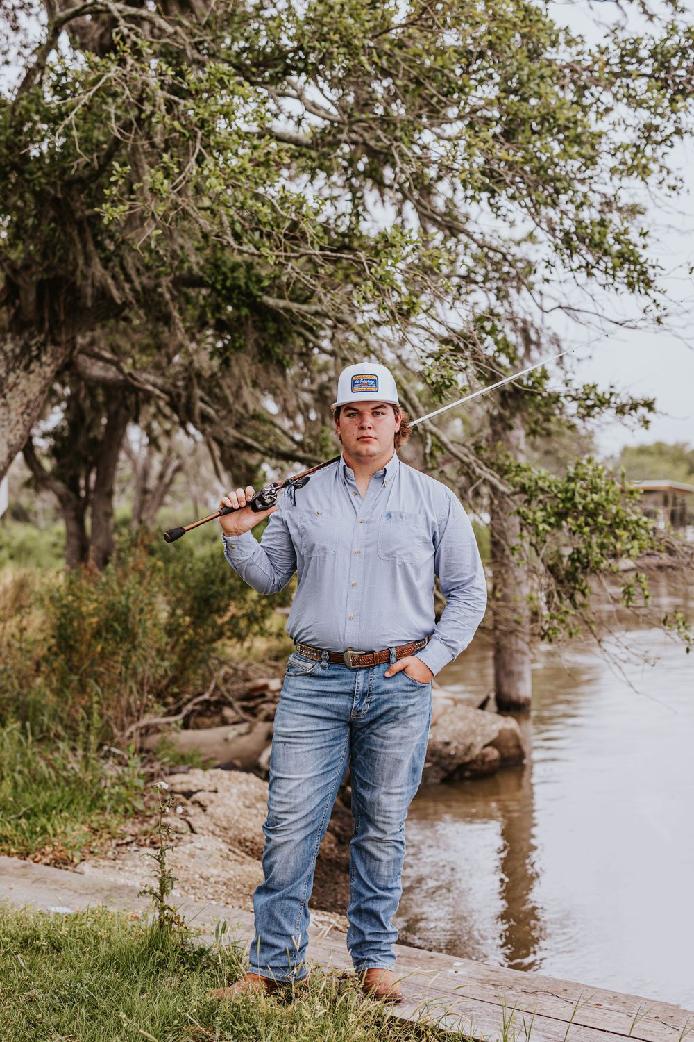 Person in light colored shirt stands near water's edge with trees in background.