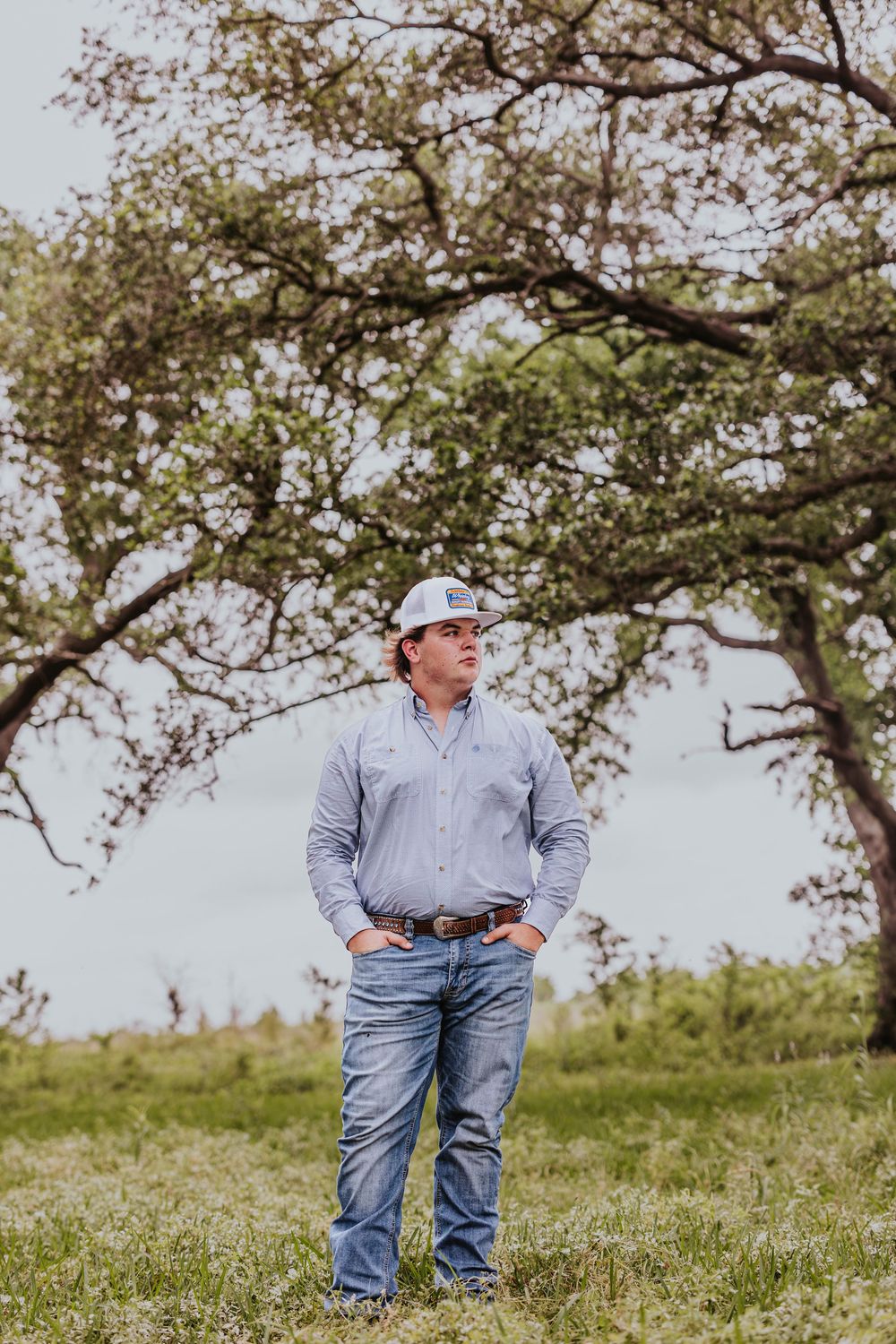Person in casual attire stands in grassy field under overhanging tree branches.