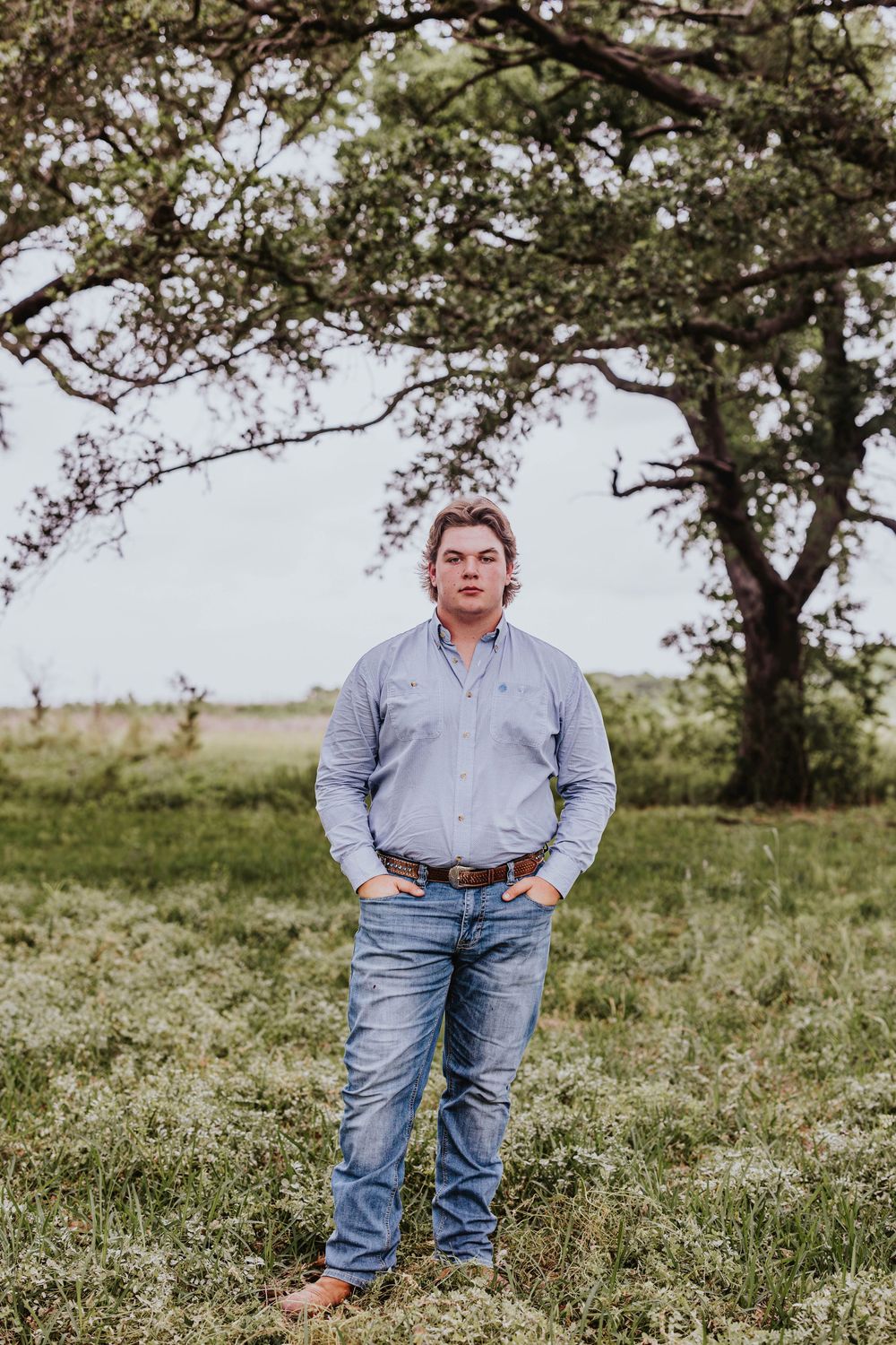 Person in light blue shirt and jeans poses in grassy field with trees in background.